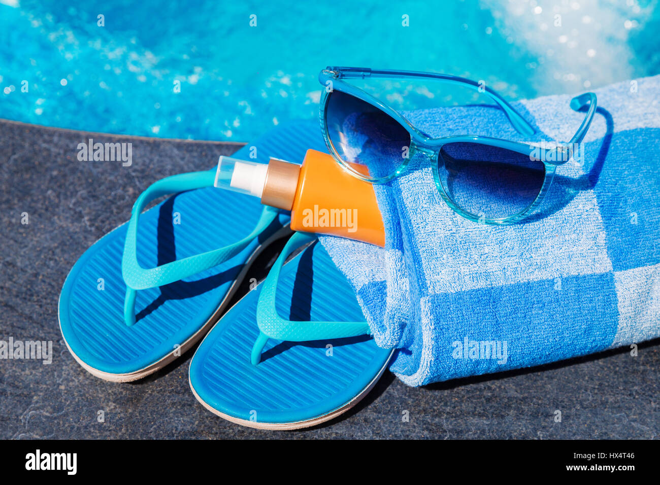 Occhiali da sole con crema di protezione solare, blu pantofole e asciugamano sul bordo di una piscina - vacanza concetto tropicale Foto Stock