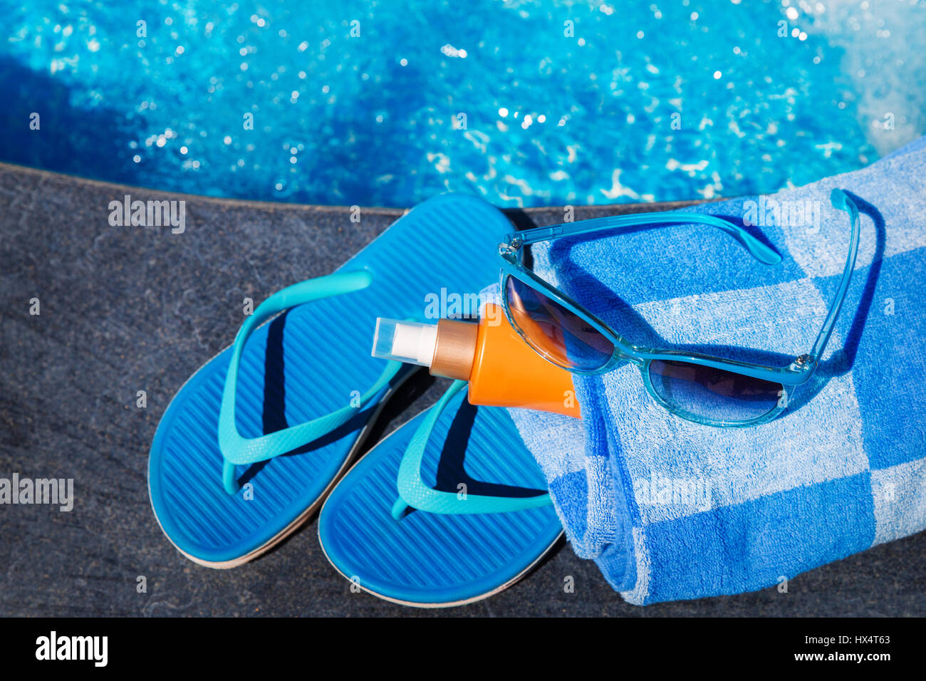 Occhiali da sole con crema di protezione solare, blu pantofole e asciugamano sul bordo di una piscina - vacanza concetto tropicale Foto Stock