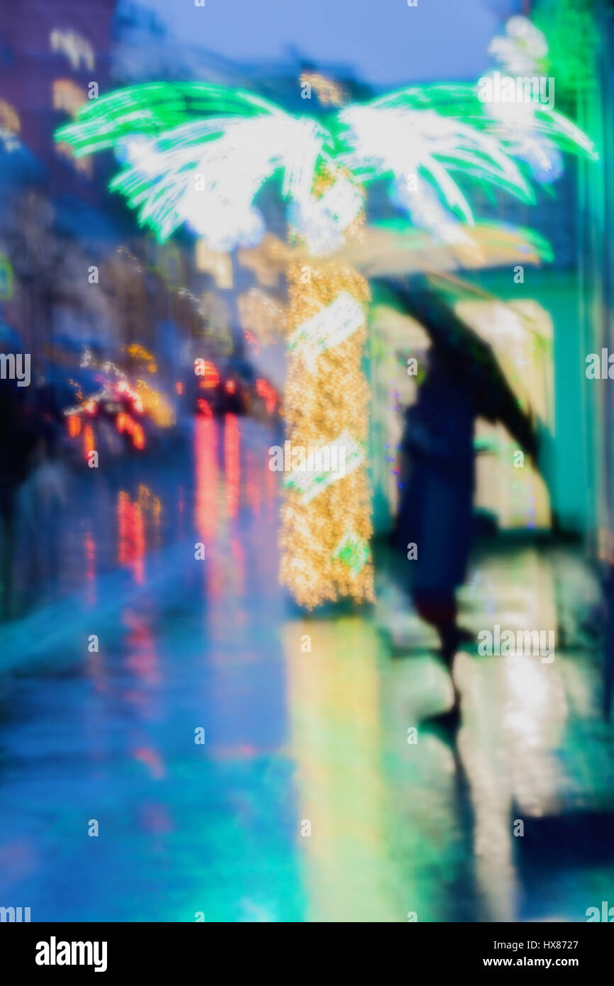 Ragazza solitaria sotto un ombrello sul marciapiede accanto a un illuminato Palm tree, strada di città sotto la pioggia, luminosi riflessi di lampioni. Intenzionale di sfocatura del movimento Foto Stock