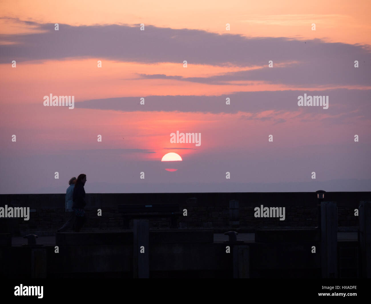 La gente a piedi passato il sole al tramonto sul molo di pietra a parete come il cloud infine si cancella e un vago ma colorato tramonto segue. Con un meteo alto insieme a seguire e temperature più calde previsto per questo fine settimana. Burton Bradstock, Dorset, Regno Unito Foto Stock