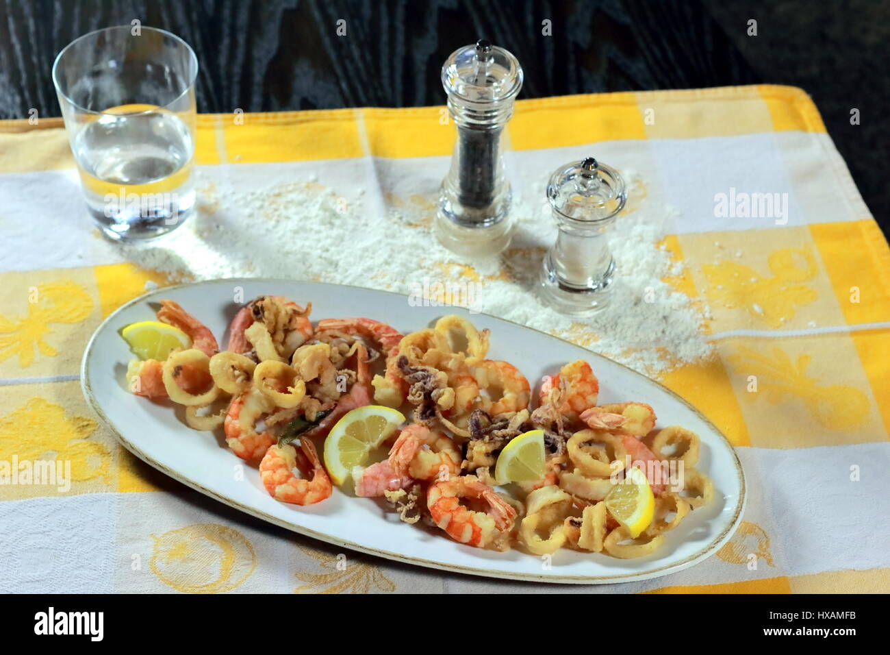 Fish Fry (pesce gamberetti - gamberetti calamari), il sale e il pepe e un bicchiere di acqua su un giallo e bianco panno da cucina giacente al buio su un tavolo di legno Foto Stock