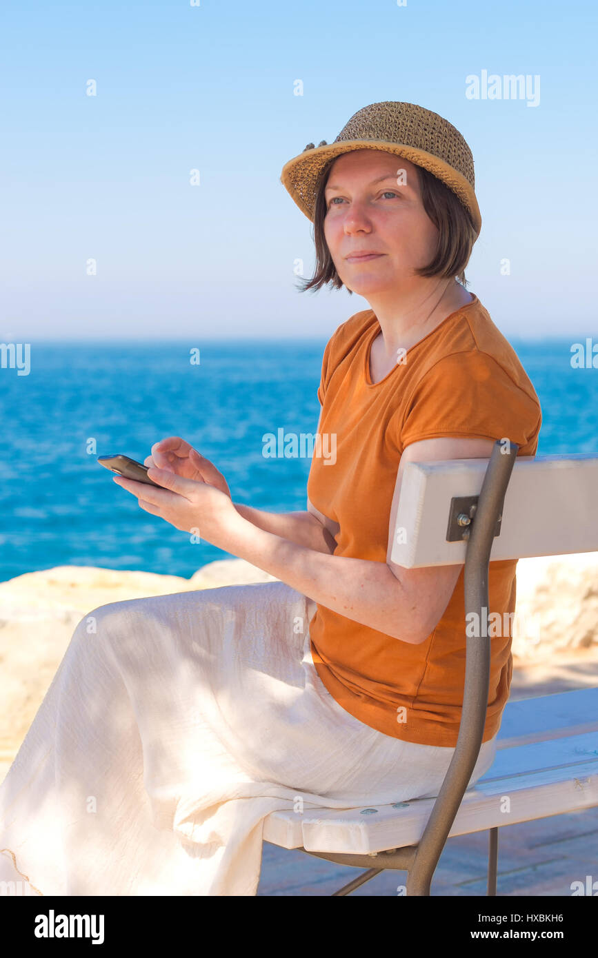 Bella adulto donna caucasica indossando Elegante cappello di paglia è utilizzando il telefono cellulare su Vacanze Estate Mare Vacanze Foto Stock