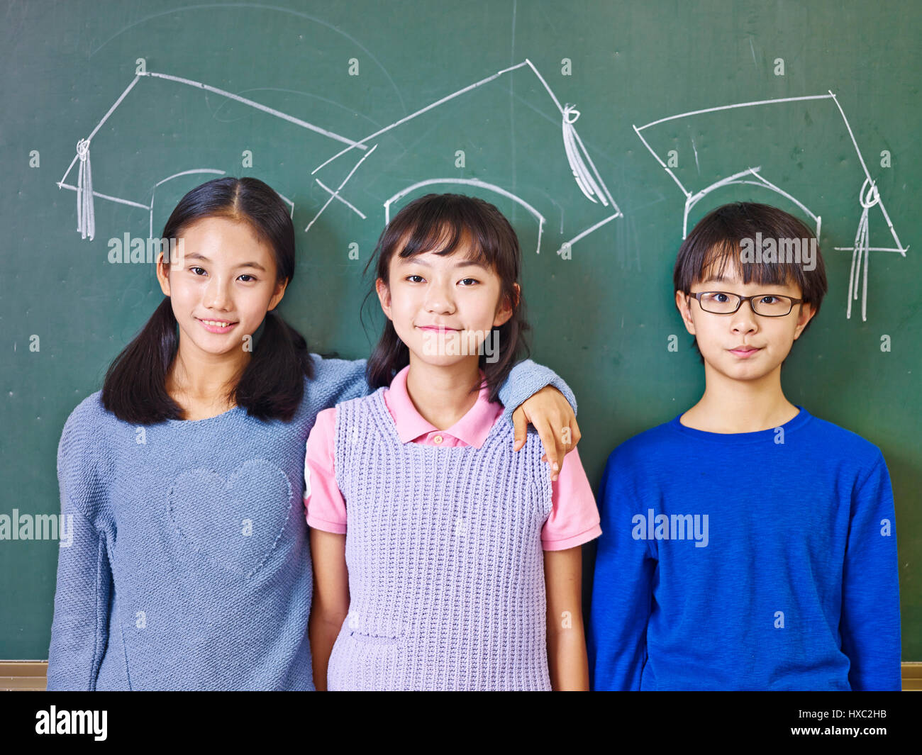 Tre asiatico i bambini delle elementari in piedi di fronte a lavagna sotto chalk-disegnato i cappucci di dottorato. Foto Stock