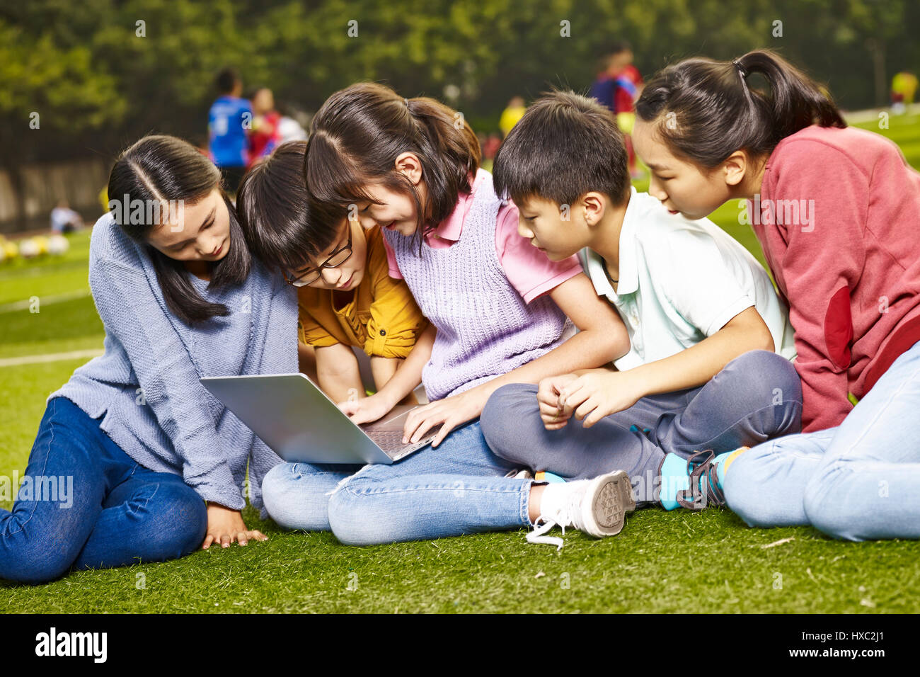 Un gruppo di asiatici scuola elementare bambini seduti su erba utilizzando laptop computer insieme. Foto Stock