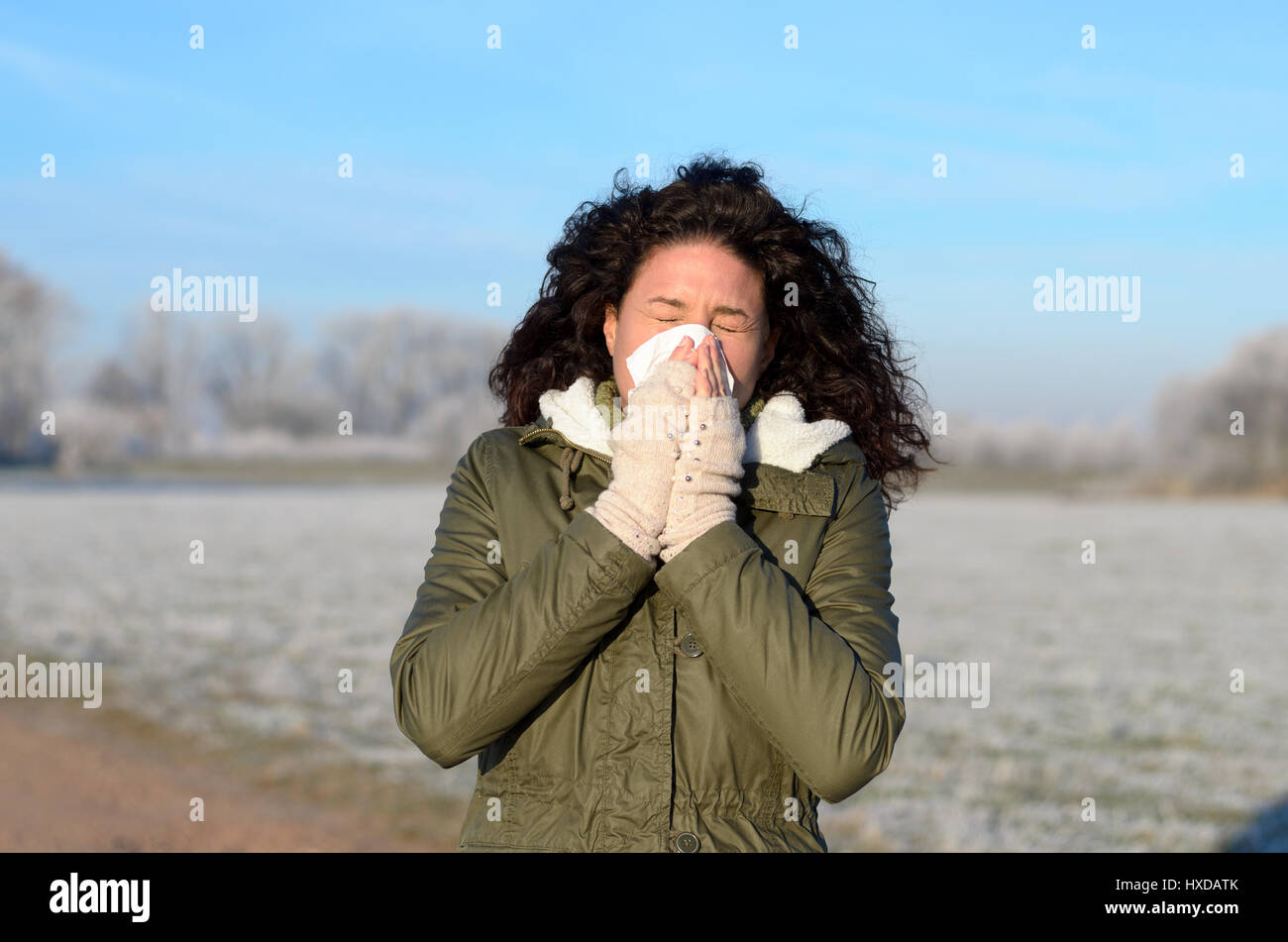 Giovane donna con l'influenza stagionale soffia il naso su un tessuto come lei gode di una passeggiata all'aperto in un freddo inverno paesaggio con la brina sui campi in un guarire Foto Stock