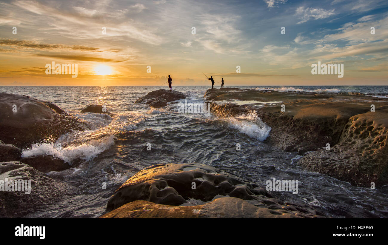 Silhouette di 3 fisherman pesca dalle rocce con le onde e il tramonto sullo sfondo. La punta del Borneo Malese. Foto Stock