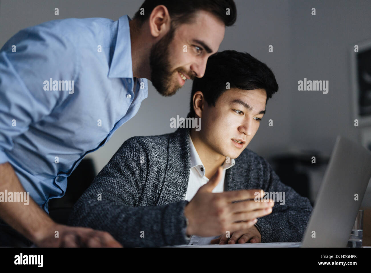 Ritratto di due lavoratori per ufficio, uno di essi asiatici, collaborando al progetto e utilizzando computer portatile in camera oscura a tarda notte, volti illuminati dalla luce della lampada Foto Stock