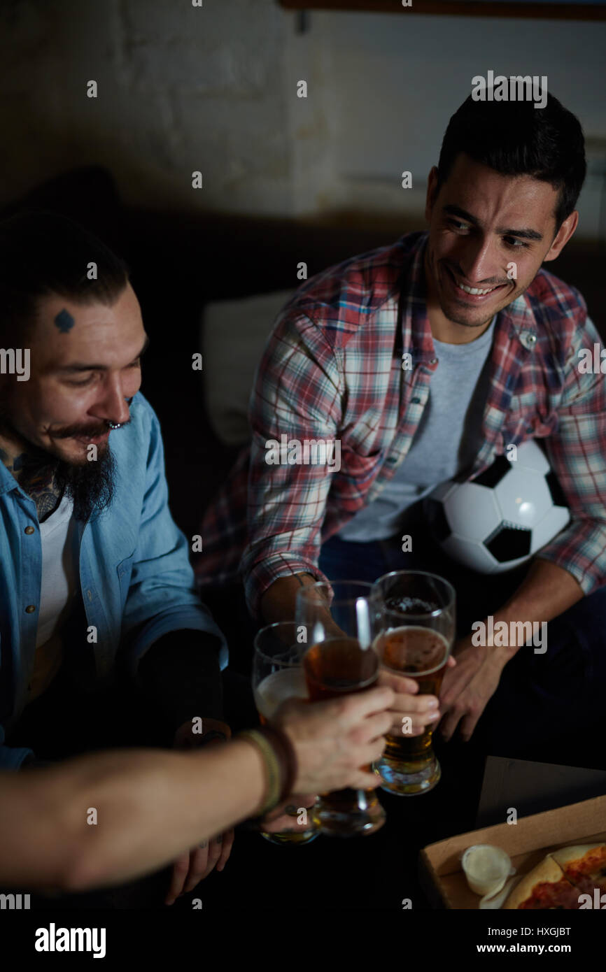 Angolo alto ritratto di gruppo di amici riuniti a casa seduti sul lettino in camera oscura la tostatura con birra e mangiare la pizza durante la visione di un tappetino di calcio Foto Stock