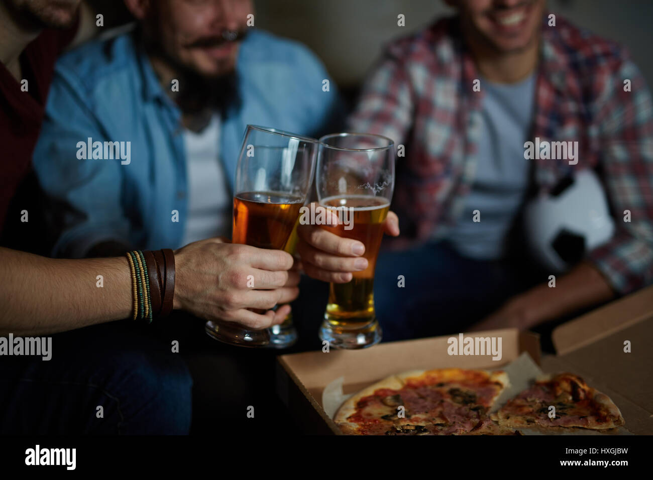 Closeup ritratto di gruppo di amici riuniti a casa seduti sul lettino in camera oscura la tostatura con bicchieri da birra e mangiare la pizza durante la visione di calcio Foto Stock