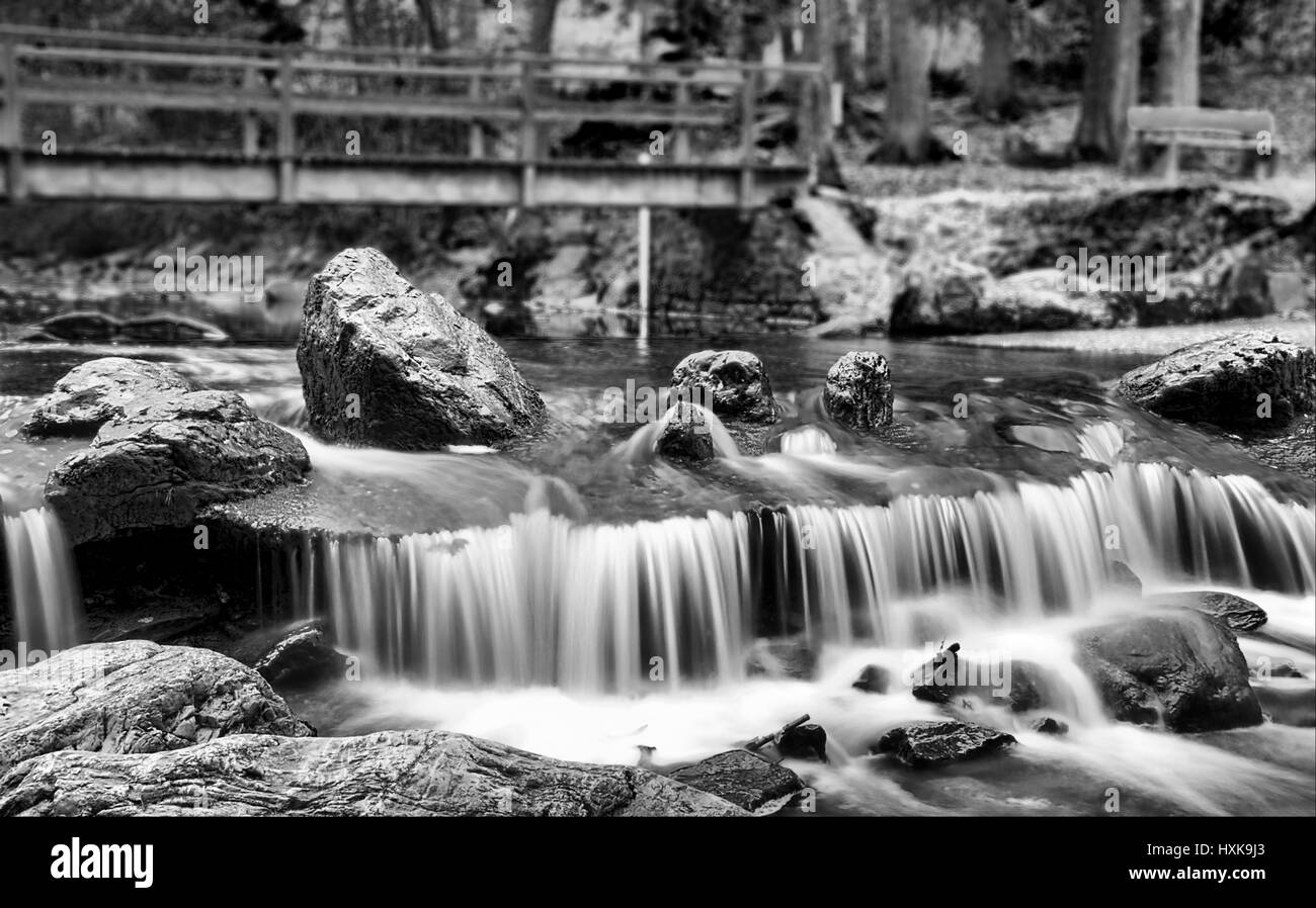 Visualizzazione bianco e nero di un basso cascata, rocce pesanti nel flusso, un ponte di legno in background. Foto Stock