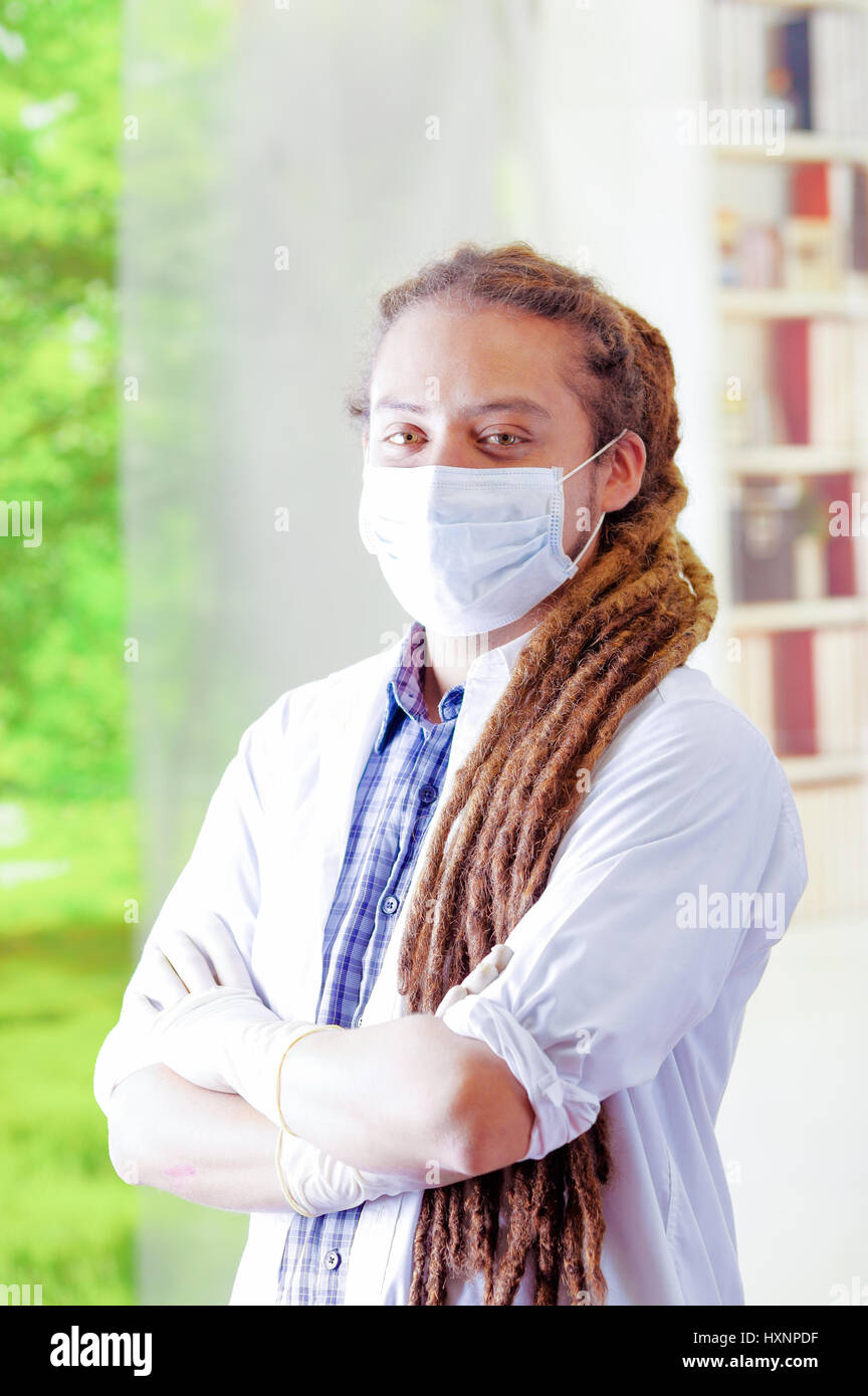 Giovane medico con lunghi dread si blocca in posa per la telecamera, indossa una maschera facciale che copre la bocca, clinica di sfondo, concetto medico Foto Stock