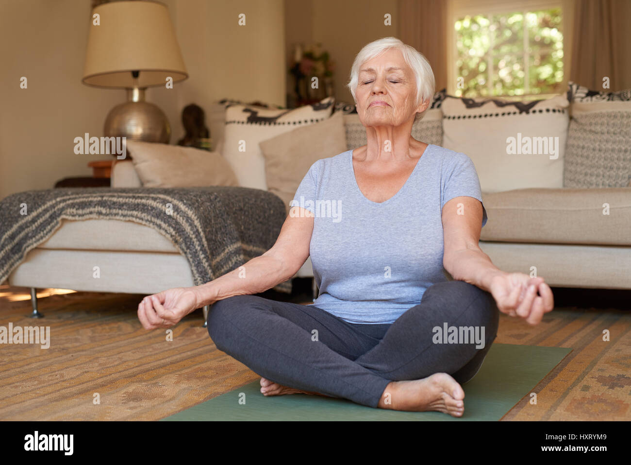 Senior donna seduta nella posizione del loto meditando a casa Foto Stock