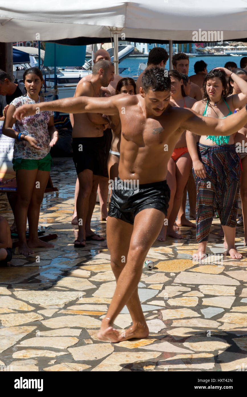 Tanzender junger Mann nel Badehose auf Dorfplatz vor Publikum in Badekleidung und ombrellone, Insel Kastellorizo, Dodekanes, Griechenland Foto Stock