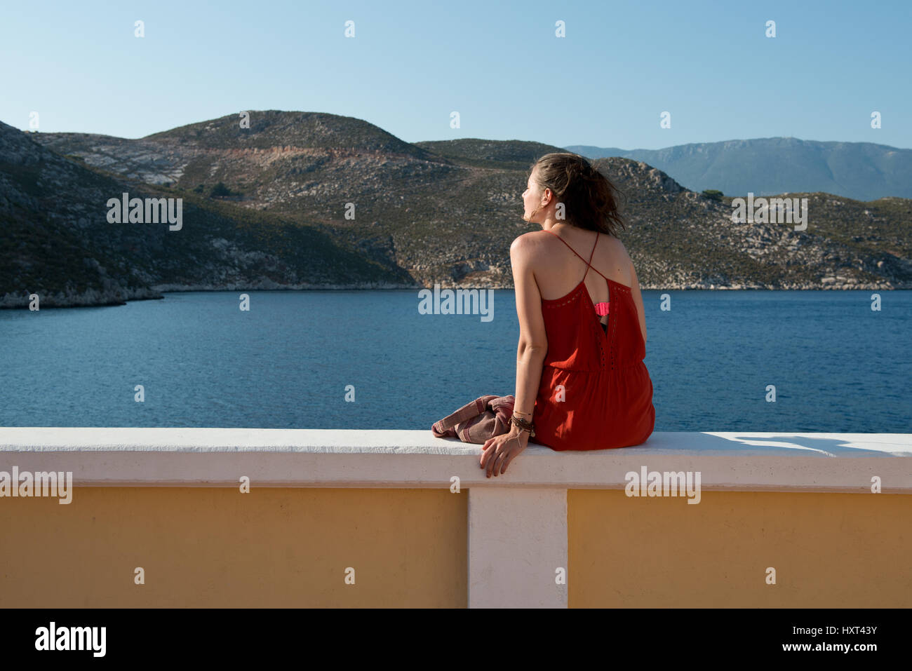 Mädchen in rotem Kleid sitzt auf Quaimauer, sieht auf Meer, dahinter kahle Hügel, Insel Kastellorizo, Dodekanes, Griechenland Foto Stock