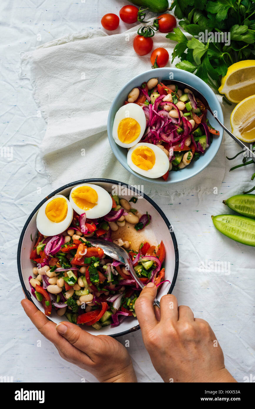 Una donna è di agitazione una pungente e colorata Bagno turco insalata di fagioli noto come piyaz in un recipiente grande e una piccola ciotola, erbe aromatiche, limone, pomodori ciliegia accompanyin Foto Stock