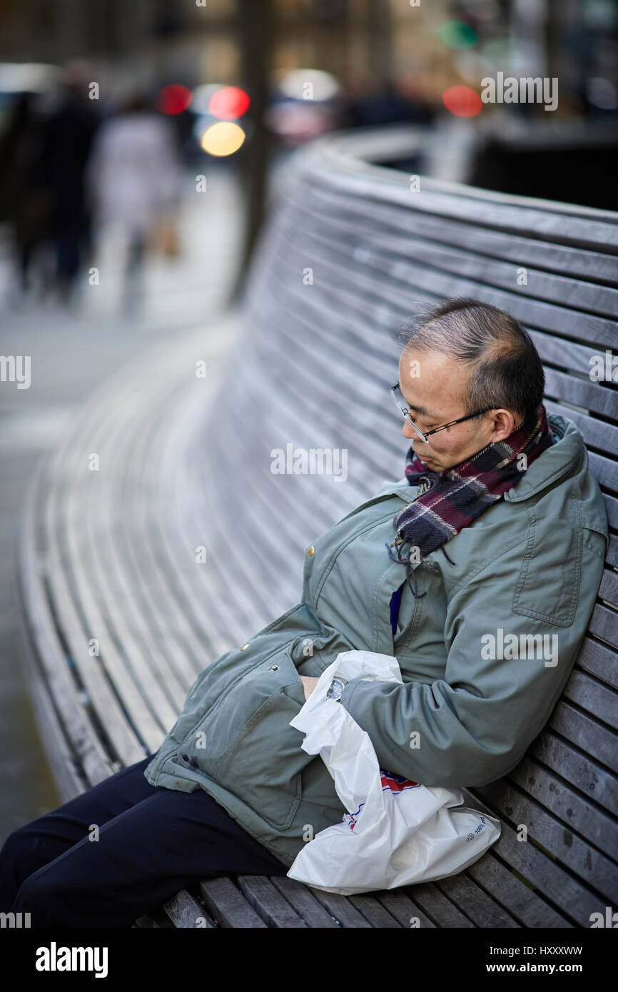 Uomo asiatico di dormire su un pubblico banco a sedere vicino a Pietro, il centro città di Manchester, Inghilterra, Regno Unito. Foto Stock