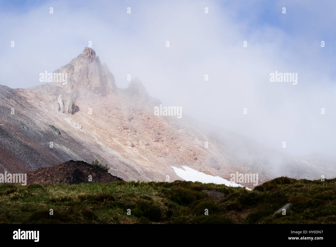 Una ripida e rocciosa Punta rossa sporge da una densa nube rotolamento sulla maggior parte della montagna. Un prato in primo piano è oscurato dalla presenza di più nuvole. Foto Stock