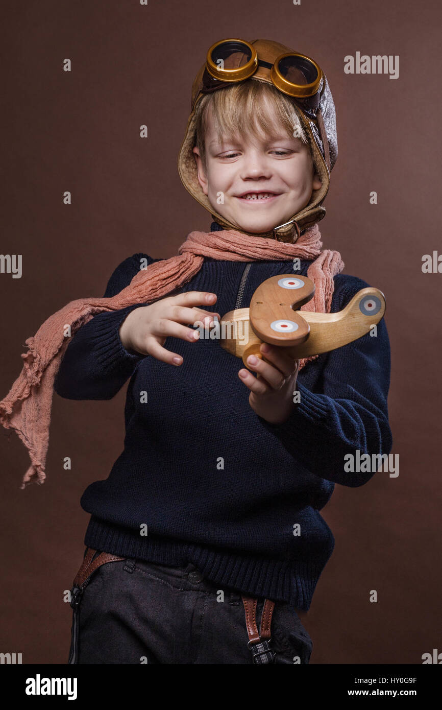Bambino felice vestito in pilot cappello e occhiali. Kid giocando con il  giocattolo di legno aereo. Sogno di libertà e di concetto. Retrò tonica.  Ritratto in studio su marrone Foto stock -