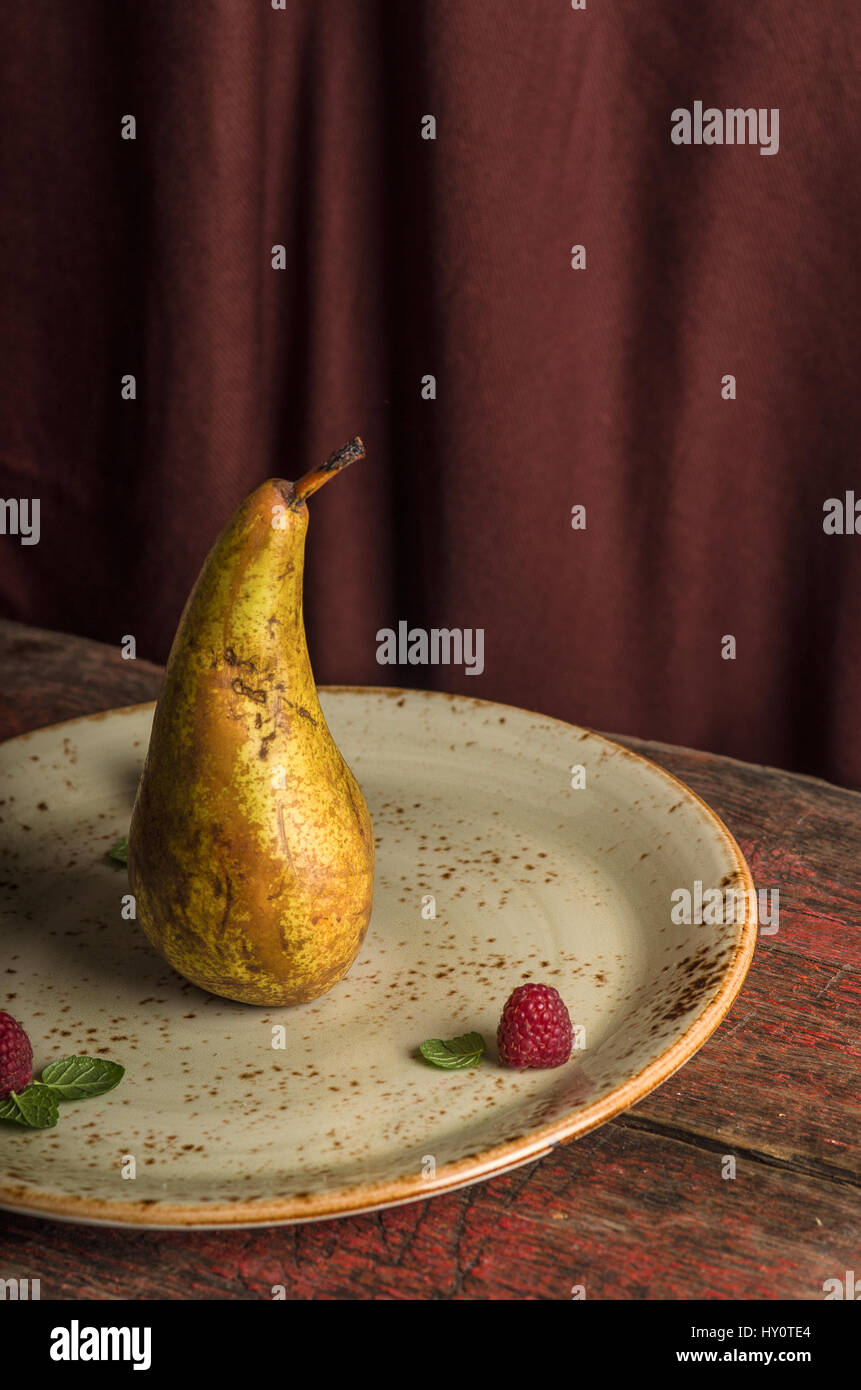 Isolato di pera di colore giallo sulla piastra con lamponi e menta su sfondo marrone Foto Stock