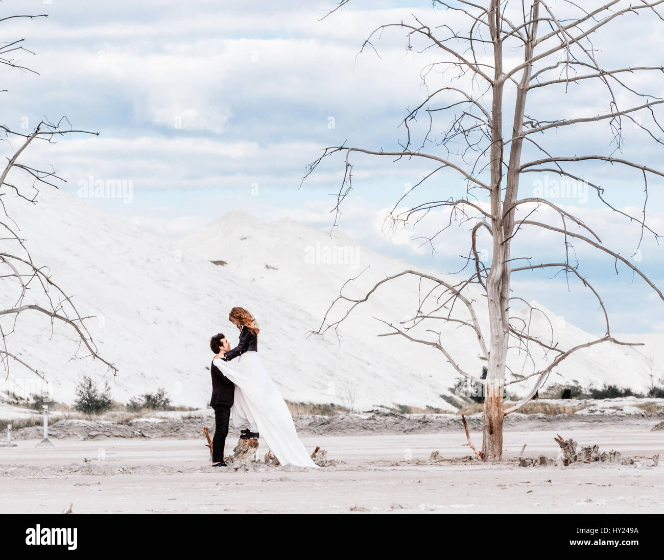Sposa con i capelli rossi si erge su essiccato fino a moncone e abbracci groom sullo sfondo del deserto con alberi appassiti, montagne di sabbia. Wedding moderna Foto Stock