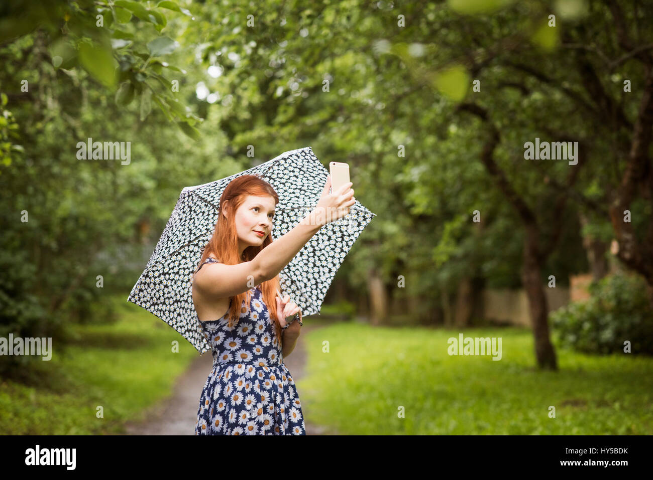 Finlandia, Pirkanmaa, Tampere, donna che indossa abiti floreali in piedi con ombrello nel parco e tenendo selfie Foto Stock