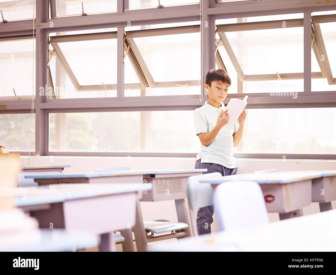 Asian schoolchild elementari in piedi da Windows per leggere un libro in aula. Foto Stock