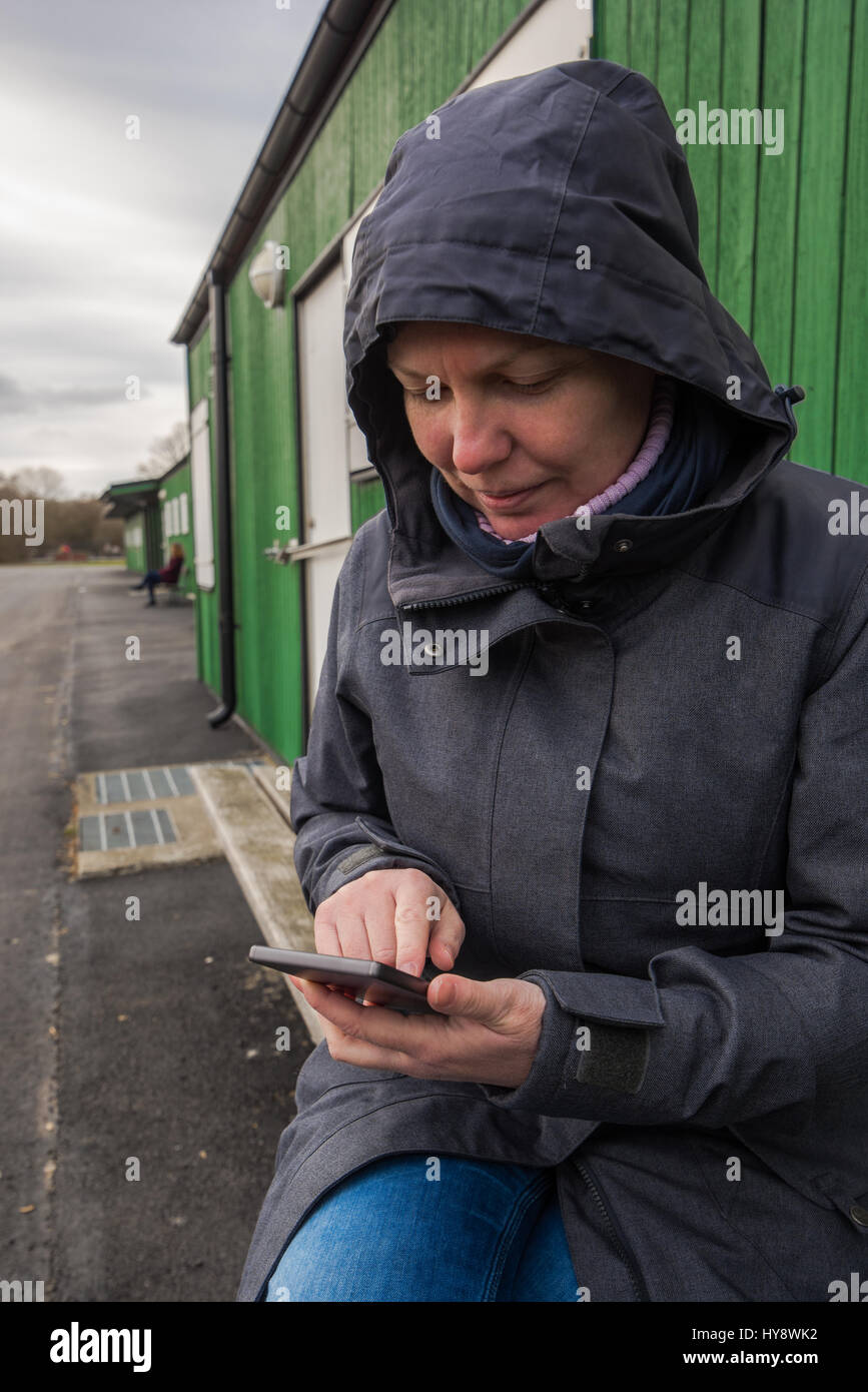 Autentica donna in giacca invernale utilizzando il telefono cellulare per tipo di messaggio sms all'aperto mentre è seduto sul banco in un freddo giorno Foto Stock