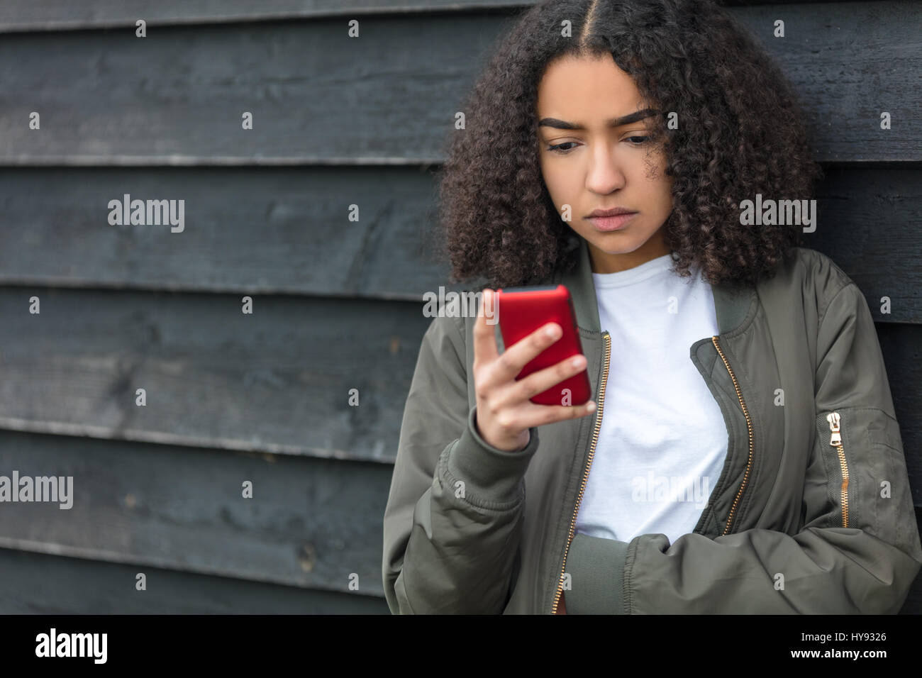 Ritratto di BELLA triste premuto razza mista americano africano ragazza adolescente femmina giovane donna texting su rosso telefono cellulare indossando bombardiere verde camicia Foto Stock