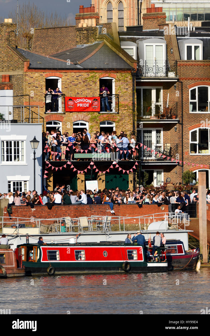 La folla di persone in attesa di guardare l'Università gara in barca sul fiume Tamigi presso Barnes, Londra. Riverside pub. I bevitori Foto Stock