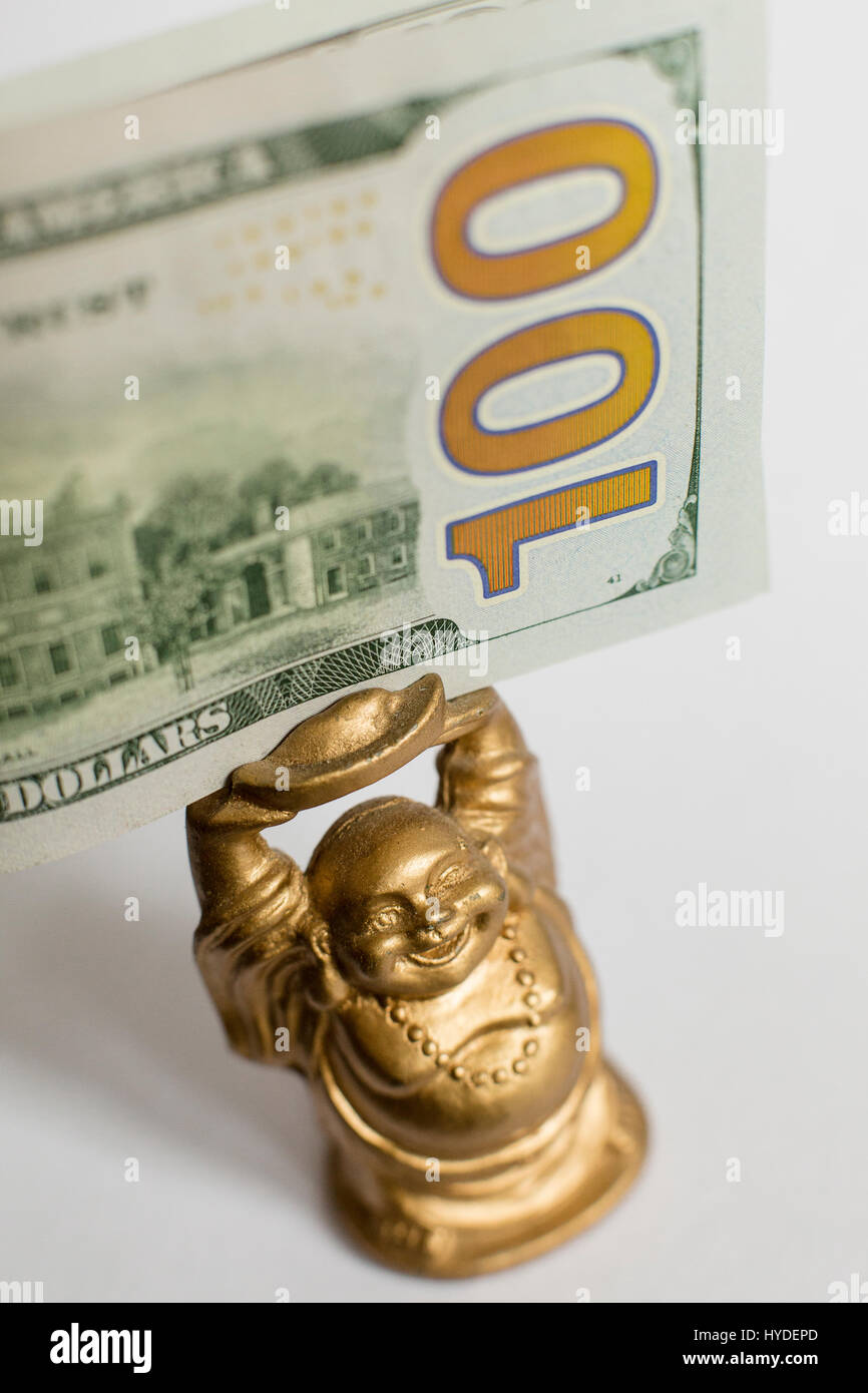 Un piccolo golden BUDDA STATUETTA in piedi sul retro bianco suolo trattiene un centinaio di dollari in valuta degli Stati Uniti sopra la sua testa Foto Stock