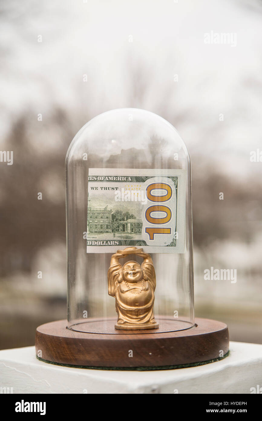 Un piccolo golden BUDDA STATUETTA in piedi in un display in vetro cupola contiene ripiegata di un centinaio di dollari sopra la sua testa Foto Stock