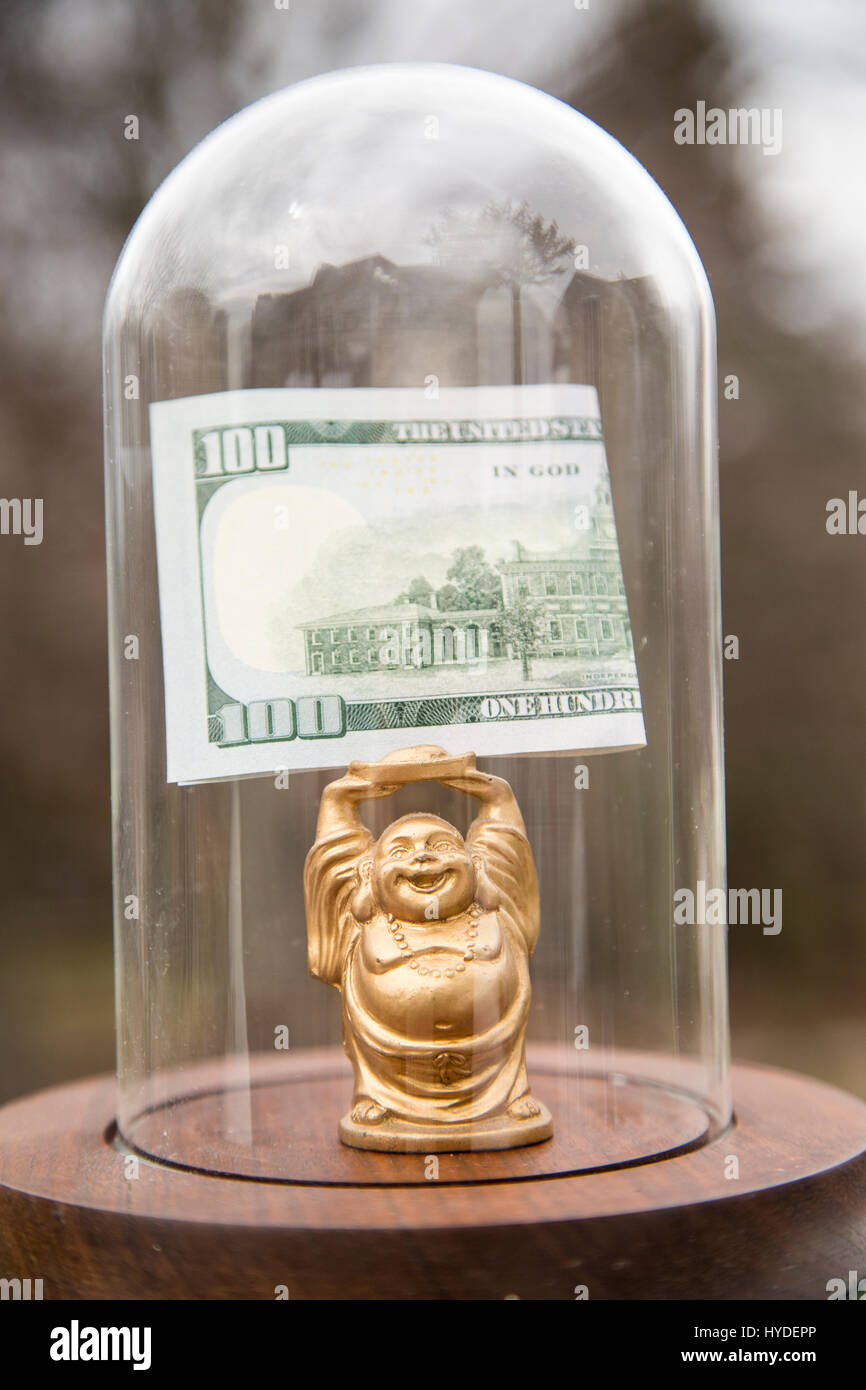 Un piccolo golden BUDDA STATUETTA in piedi in un display in vetro cupola contiene ripiegata di un centinaio di dollari sopra la sua testa Foto Stock
