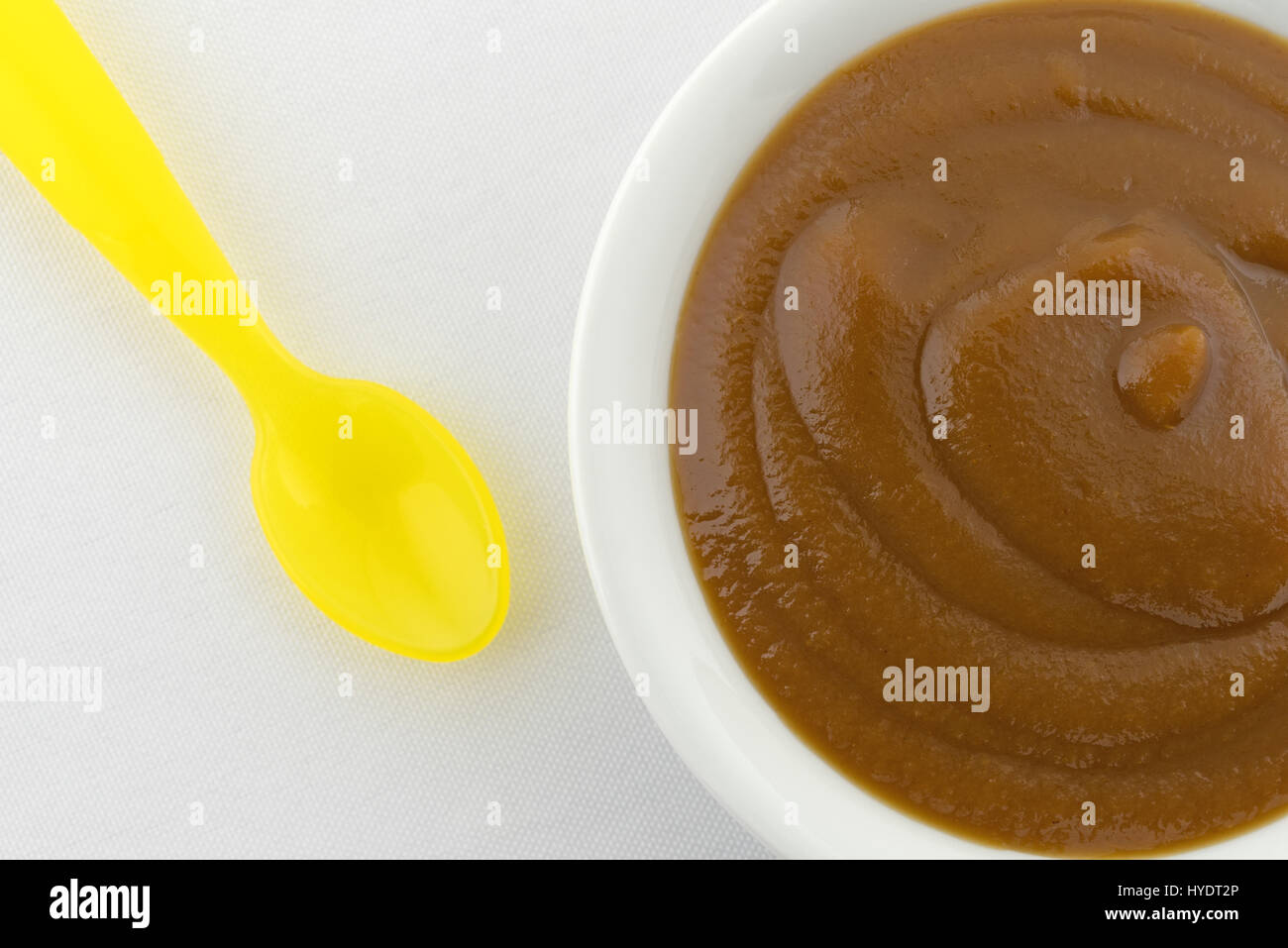 Top vista ravvicinata di una piccola ciotola riempita con Apple e potare la purea di pappe per bambini più un giallo cucchiaio di plastica a lato sulla cima di una tovaglia bianca. Foto Stock