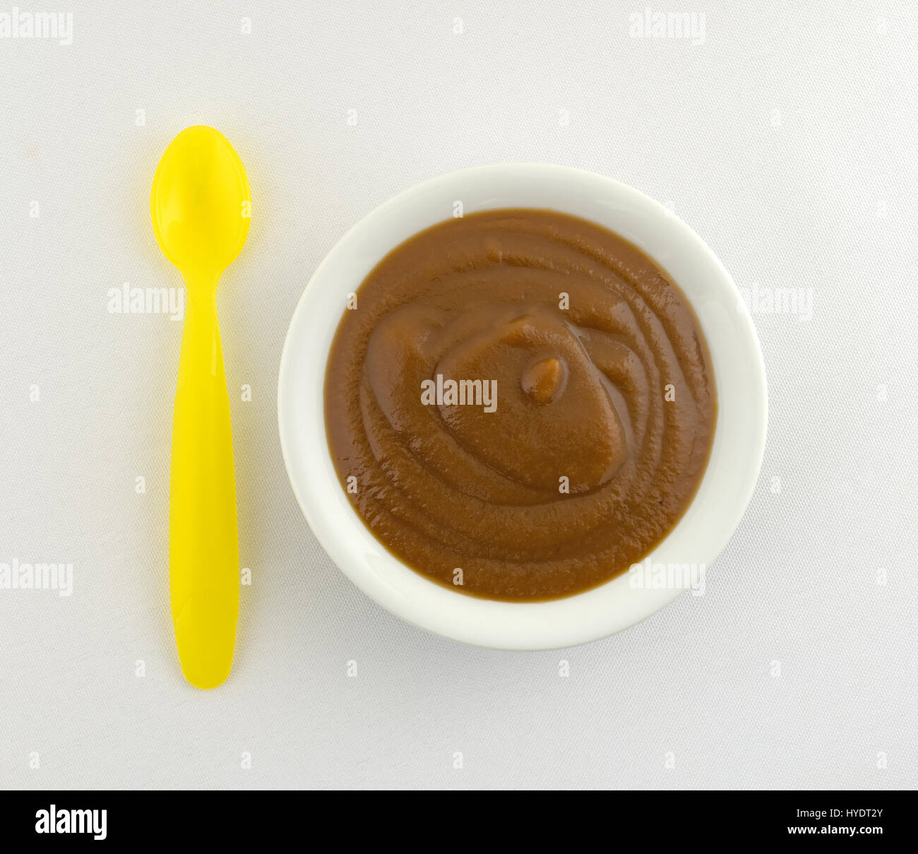 Vista dall'alto di una piccola ciotola riempita con Apple e potare la purea di pappe per bambini più un giallo cucchiaio di plastica a lato sulla cima di una tovaglia bianca. Foto Stock