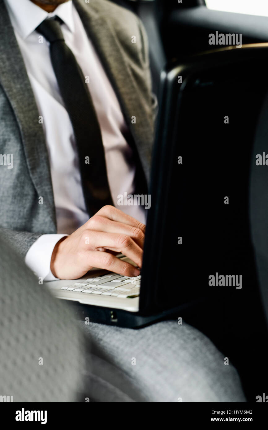 Primo piano di un giovane imprenditore caucasico in un abito grigio usando un computer portatile seduta sul sedile posteriore di una macchina Foto Stock