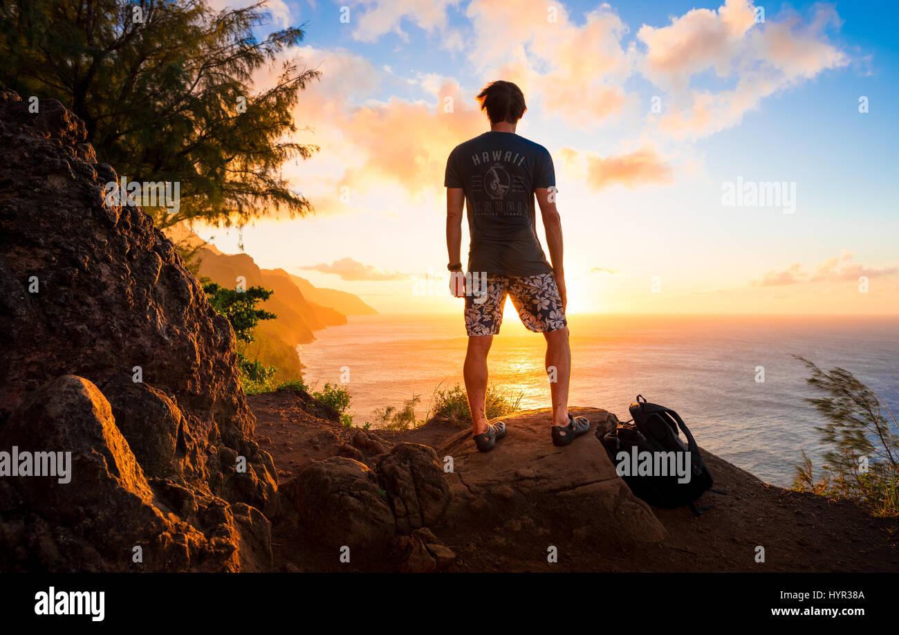 Un maschio di escursionista guarda il tramonto su un Kauii sentiero escursionistico che si affaccia su un tramonto sull'oceano Pacifico. Foto Stock