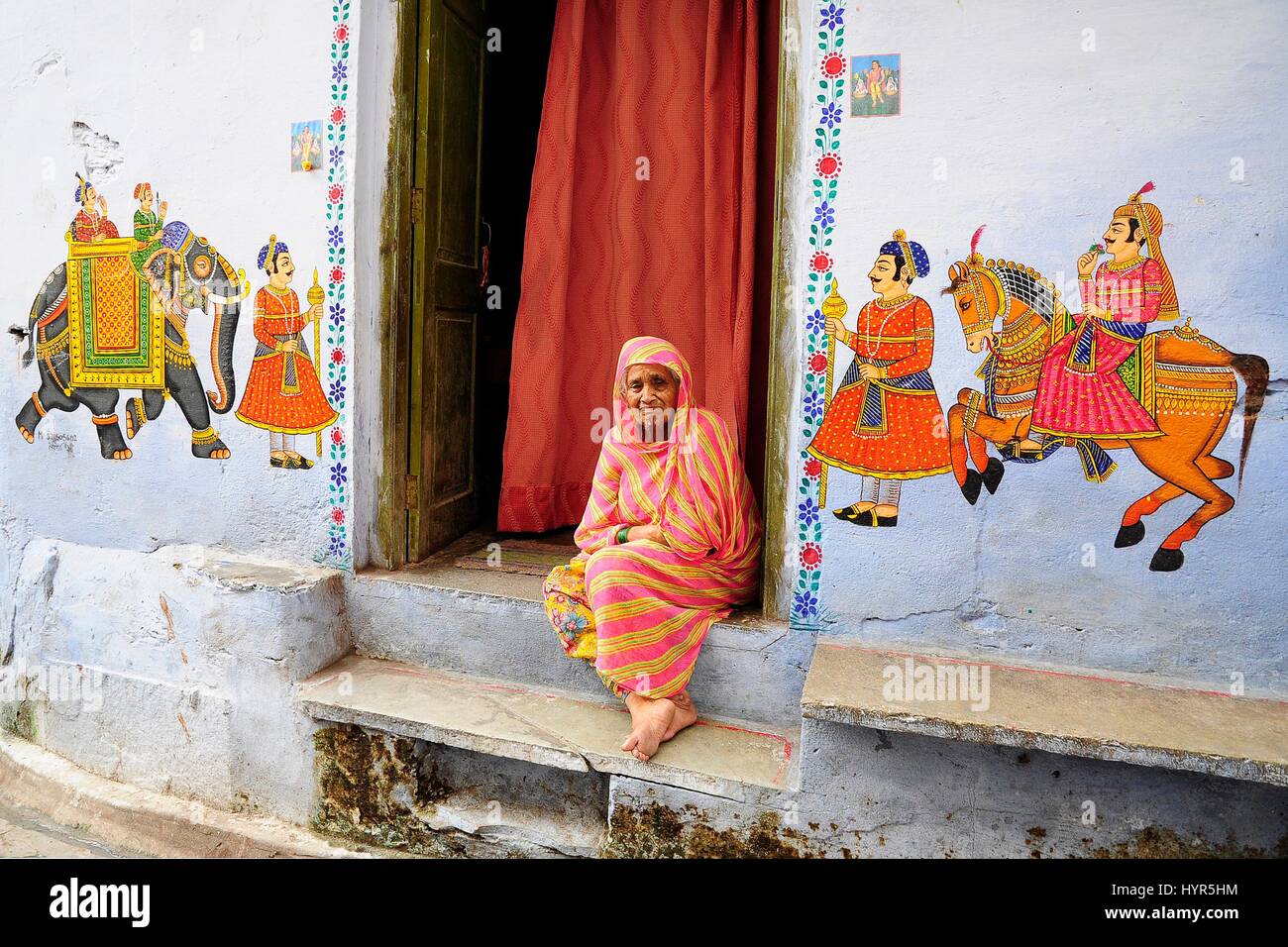 Udaipur, India, settembre 12, 2010: vecchia donna indiana seduto di fronte a lei huse su un terreno con dipinti su una parete. Foto Stock