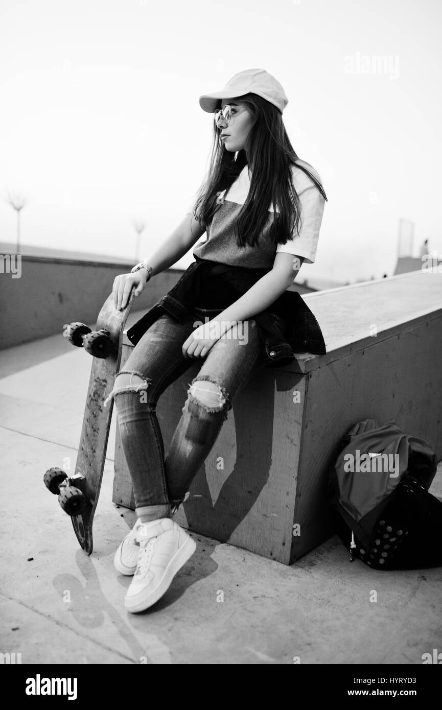 I giovani adolescenti ragazza urbano con lo skateboard, usura sui bicchieri, cappuccio e jeans strappati a skate park la sera. Foto Stock