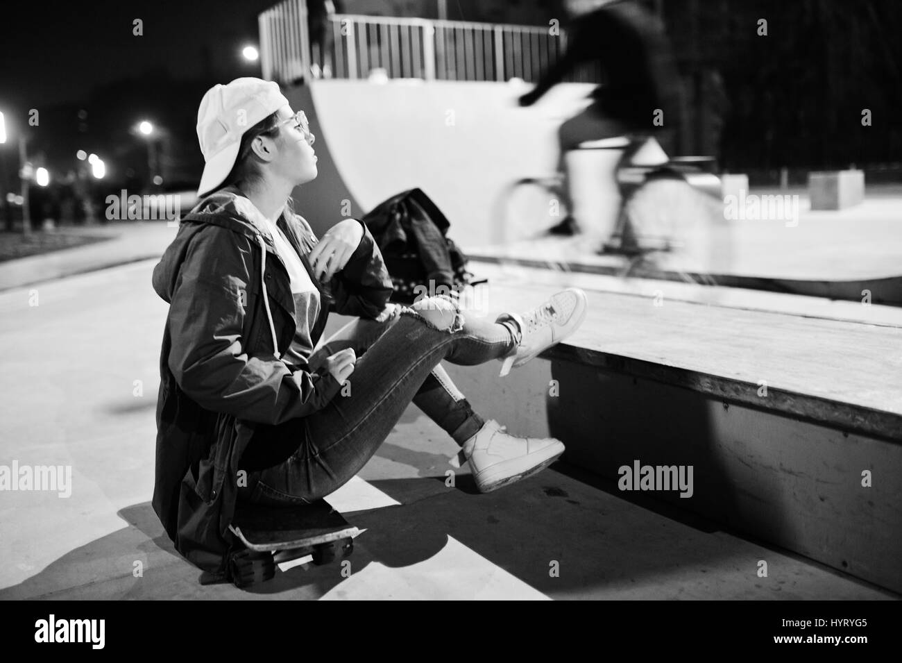 I giovani adolescenti ragazza urbano con lo skateboard, usura sui bicchieri, cappuccio e jeans strappati a skate park la sera. Foto Stock