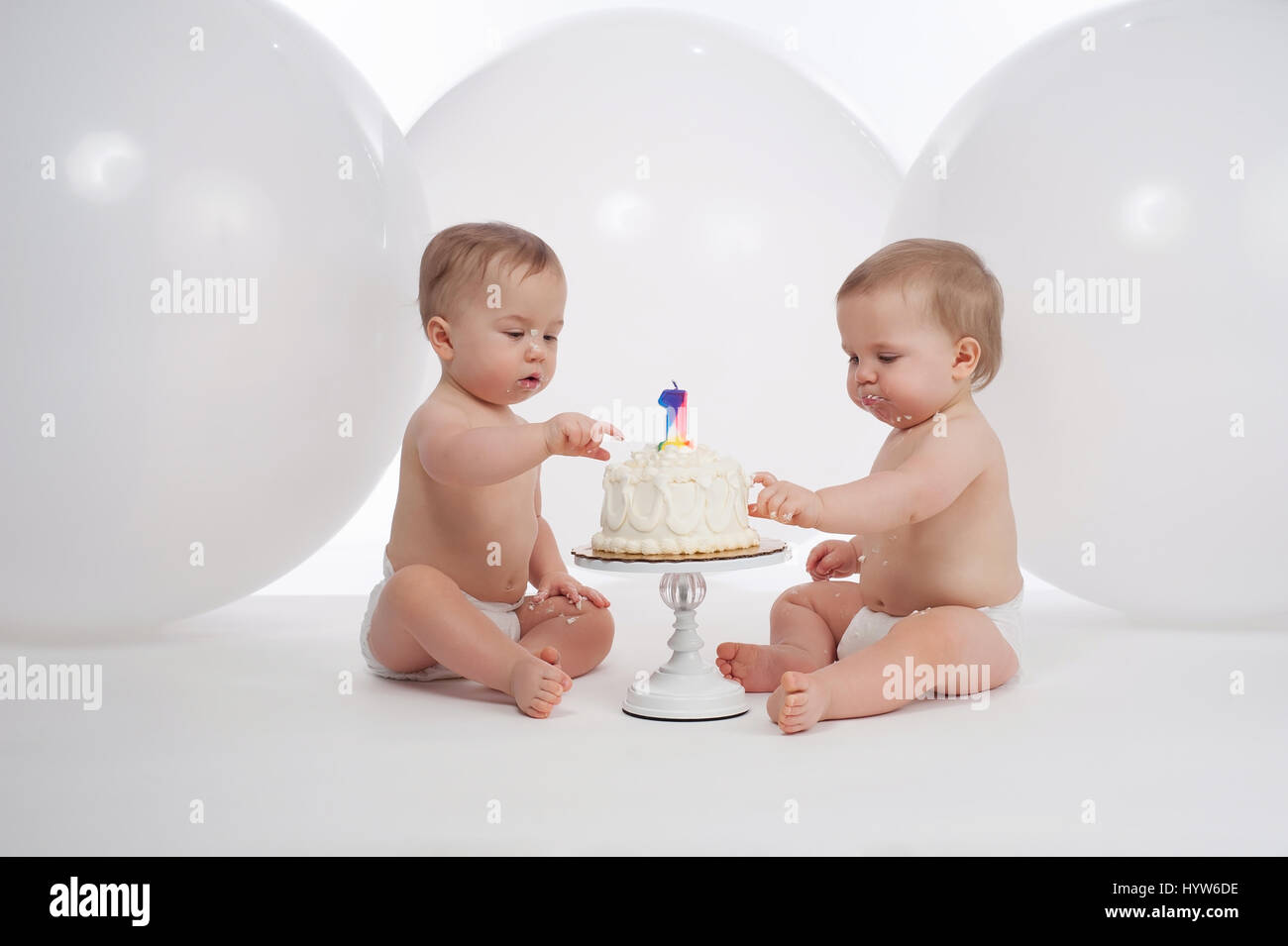 Un anno di età i fratelli gemelli che indossa pannolini e mangiare la loro torta di compleanno. Girato in studio con grandi, palloncini bianchi in background. Foto Stock