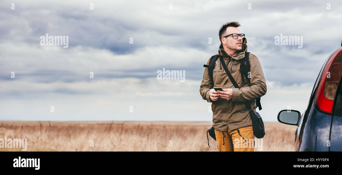 Escursionista in piedi nella prateria dall'auto utilizzando smart phone e guardando a distanza Foto Stock