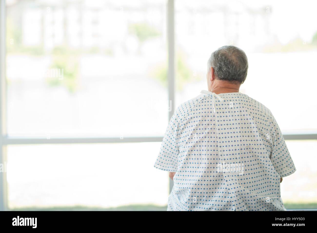 Paziente di sesso maschile che indossa camice guardando attraverso la finestra dell'ospedale, in vista posteriore Foto Stock