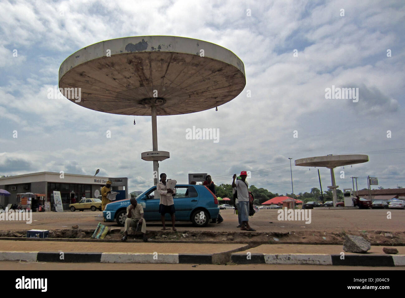 Auto blu e un po' di persone davanti a una vecchia stazione di benzina in attesa di fare rifornimento, accanto a una strada nella città di Lagos, Nigeria Foto Stock