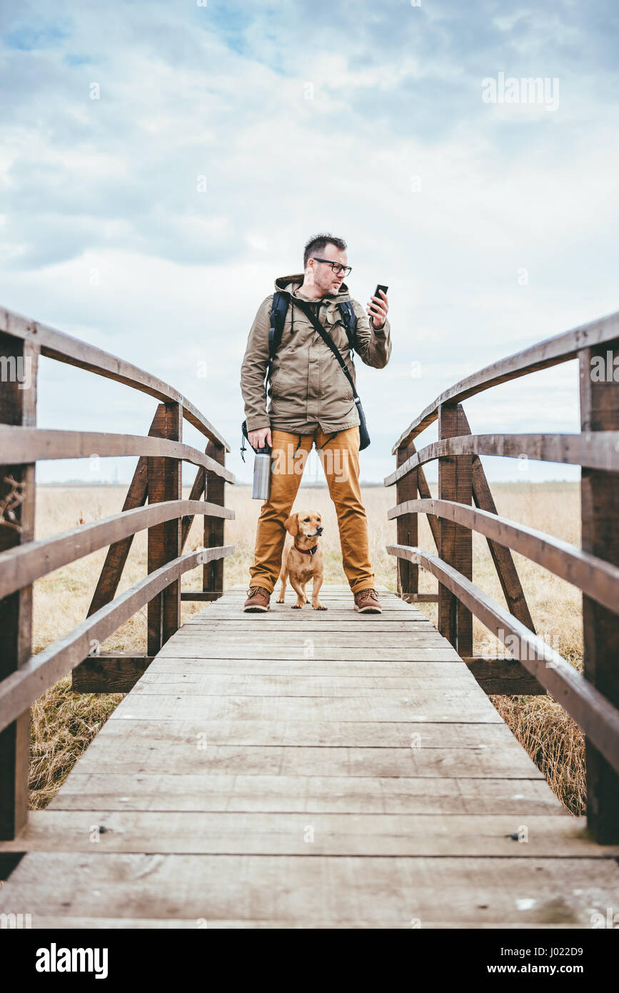 Escursionista in piedi su un ponte di legno con il suo cane e utilizzo di smart phone Foto Stock