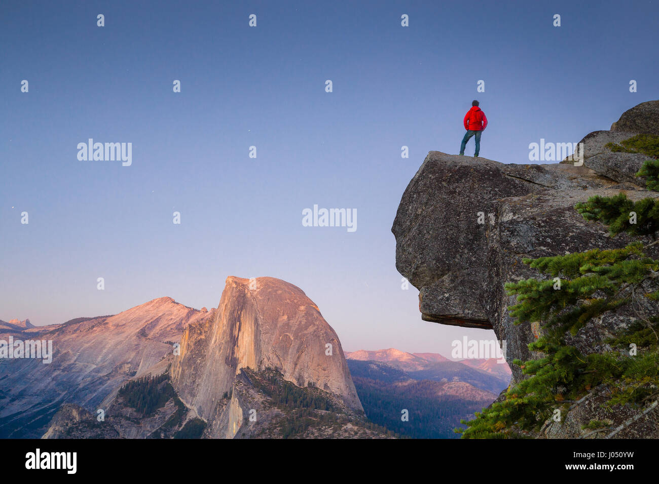 Un intrepido escursionista è in piedi su una roccia a strapiombo godendo della vista verso il famoso Half Dome presso il Glacier Point si affacciano al tramonto, Yosemite NP, STATI UNITI D'AMERICA Foto Stock