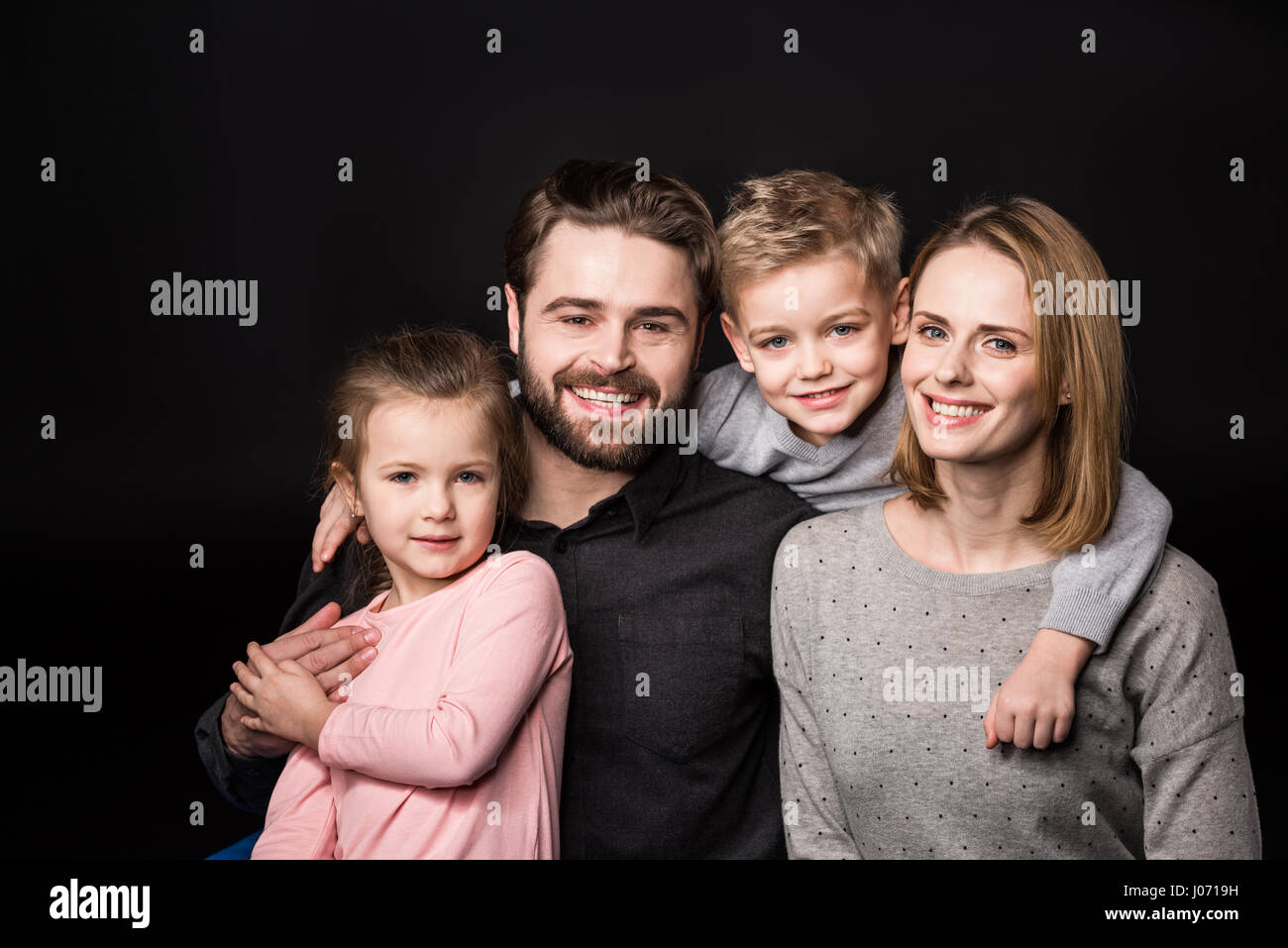 Felice giovane famiglia con due bambini standing abbracciando e sorridente in telecamera su nero Foto Stock