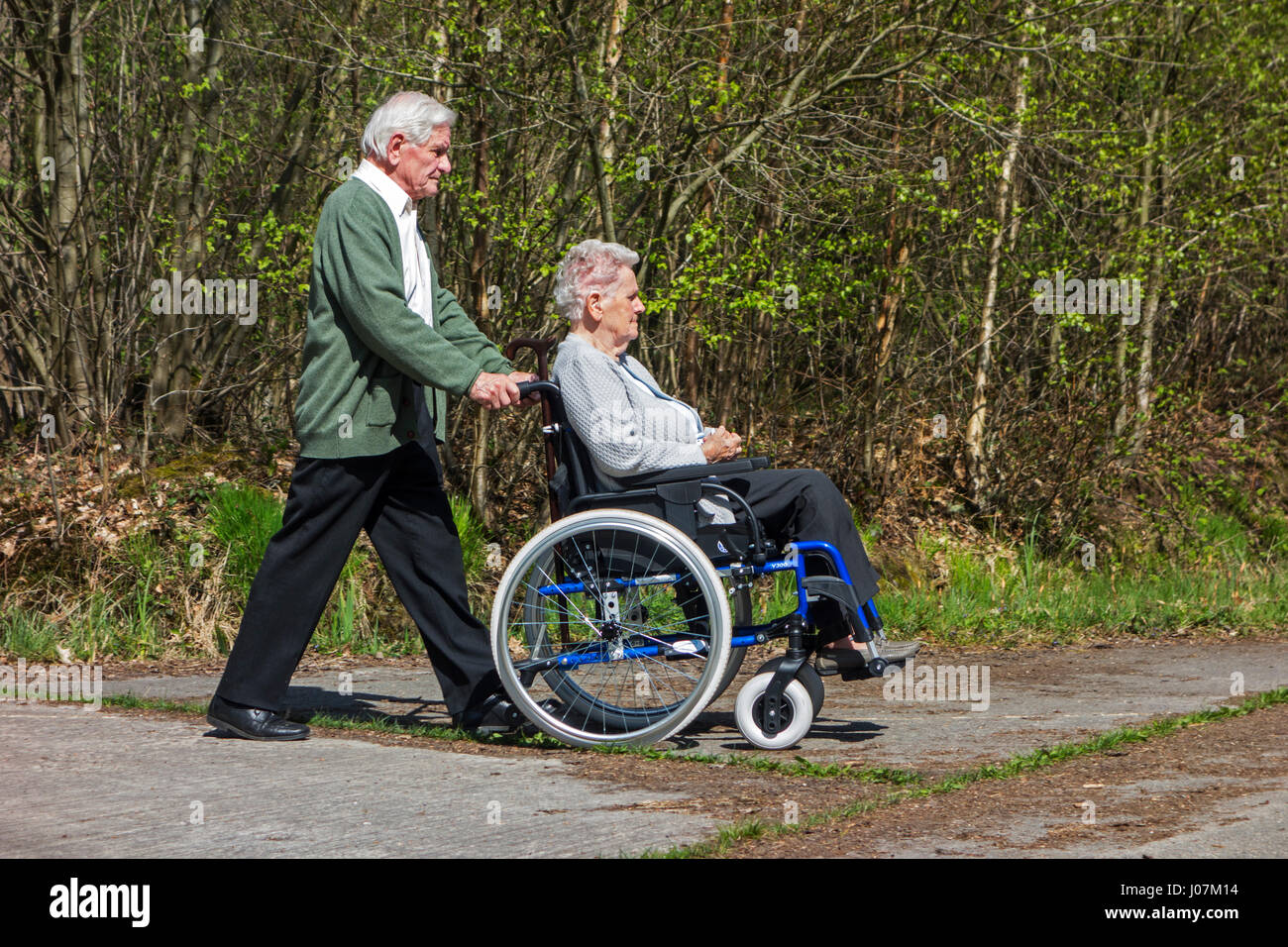 Ritirato il marito prendendo anziani disabili moglie in carrozzella per una passeggiata nel parco in una giornata di sole in primavera Foto Stock