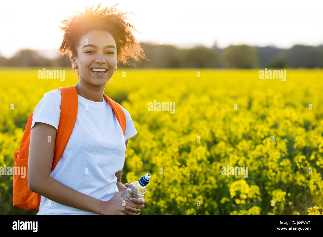 Outdoor ritratto della bella felice razza mista americano africano ragazza adolescente femmine giovane donna con una bottiglia d'acqua escursionismo con back pack in un campo di Foto Stock