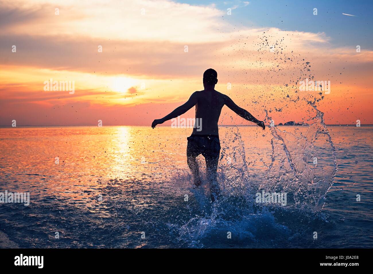 Gioiosa giovane uomo che corre in acqua di mare tropicale durante il bellissimo tramonto. Foto Stock