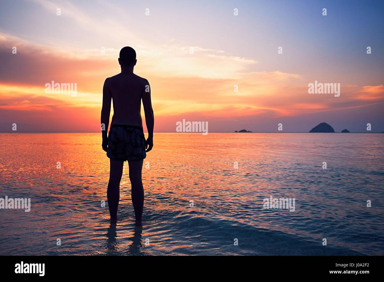 Pensieroso giovane uomo in piedi in acqua del mare tropicale durante il bellissimo tramonto. Foto Stock